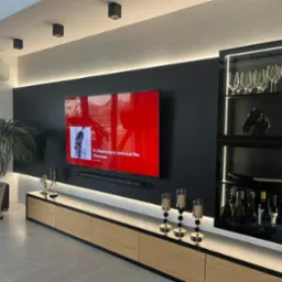 Nowoczesny salon z podświetlaną szafką RTV z drewnianymi frontami, czarną ścianą TV i przeszkloną szafką na alkohol, całość dopełniona dekoracjami i rośliną doniczkową.