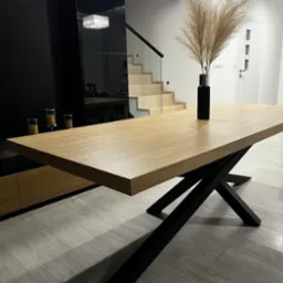 Blat drewniany stołu jadalnego z nogami w kształcie litery X, w nowoczesnym wnętrzu z czarną szafką i schodami w tle.