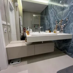 Nowoczesna łazienka z białą, wiszącą umywalką i szafką w odcieniach beżu, marmurowa ściana w odcieniach szarości i błękitu, lustro odbijające wnętrze łazienki i osobę robiącą zdjęcie telefonem.