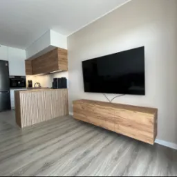 Nowoczesne wnętrze z drewnianą zabudową kuchenną, wyspą z pionowych listew, wiszącą szafką RTV z forniru i dużym telewizorem na ścianie.