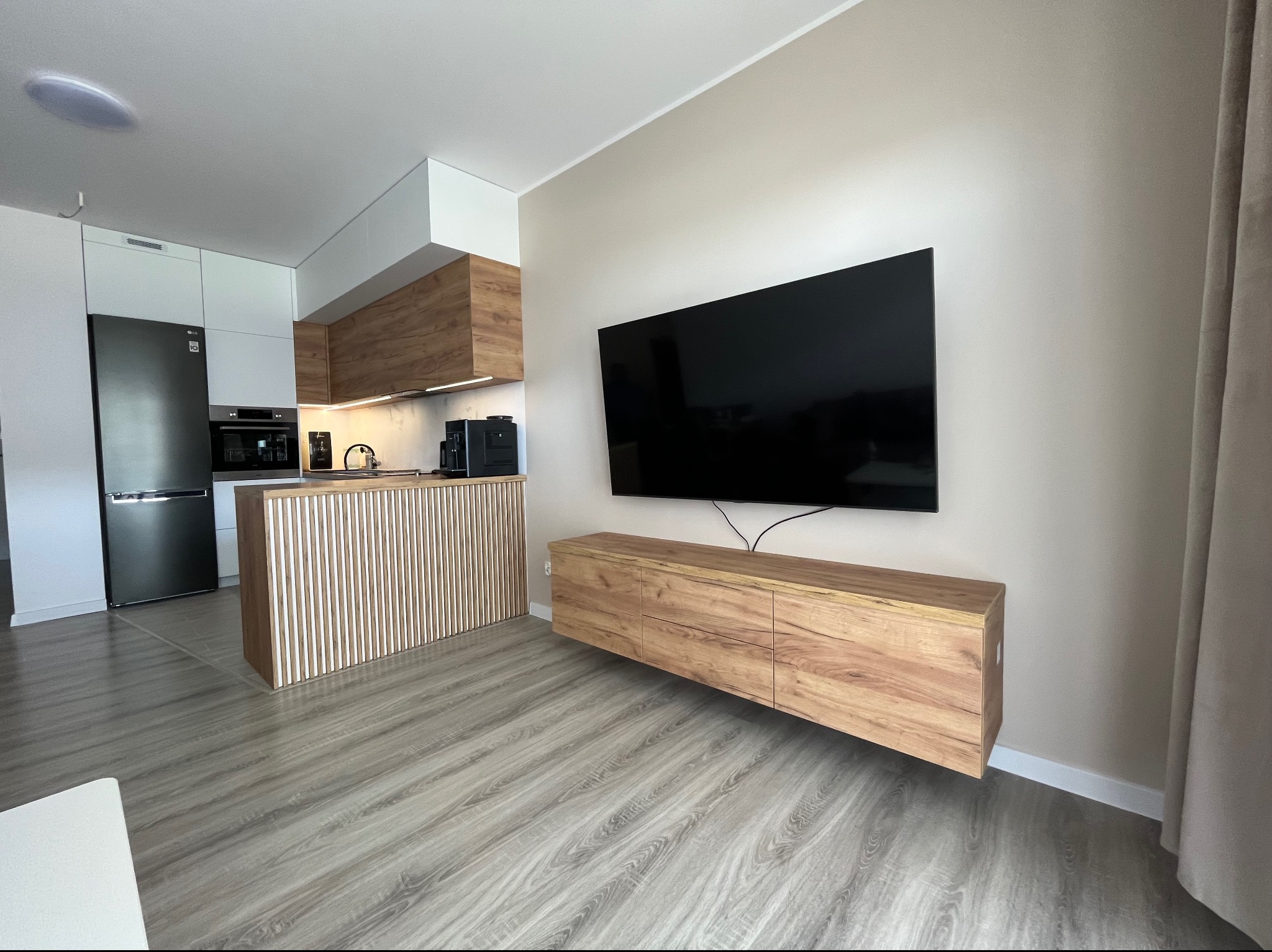 Nowoczesne wnętrze z drewnianą zabudową kuchenną, wyspą z pionowych listew, wiszącą szafką RTV z forniru i dużym telewizorem na ścianie.