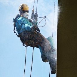 High five usługi alpinistyczne - Pracownik w uprzęży, z maską i kasku, czyści elewację budynku pod ciśnieniem. Widoczne liny, dysza i strumień wody na tle błękitnego nieba.