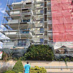 High five usługi alpinistyczne - Budynek w rusztowaniach, zabezpieczony siatką, przygotowywany do prac elewacyjnych. Widoczny pracownik w stroju roboczym. Ujęcie z dołu, w tle błękitne niebo.