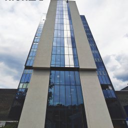 High five usługi alpinistyczne - Wysoki budynek biurowy z przeszkloną fasadą, na której pracownik na linach wykonuje prace na wysokości. Widok z dołu, pochmurne niebo w tle.