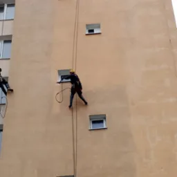 Dwóch pracowników na linach czyści elewację wysokiego budynku za pomocą myjek ciśnieniowych, widoczne różnice w kolorze elewacji przed i po czyszczeniu.