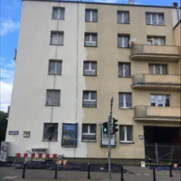 Malowanie elewacji bloku mieszkalnego w trakcie realizacji, widoczne częściowo pomalowane fragmenty, zabezpieczone okna, robotnik w oknie, farby i taśmy ostrzegawcze na chodniku.