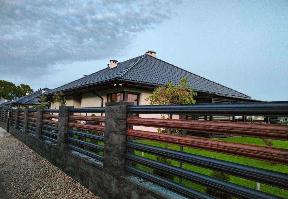 Nowoczesne ogrodzenie z elementami betonowymi i poziomymi przęsłami w kolorze czarnym i drewnopodobnym, na tle domu z ciemną dachówką i pochmurnego nieba.