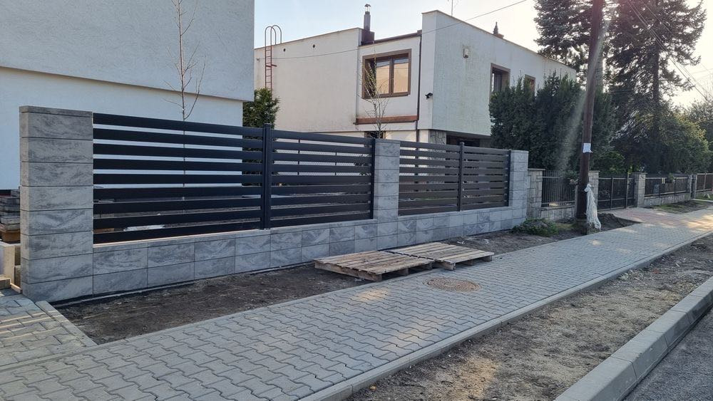 Nowoczesne ogrodzenie z betonowymi słupkami i poziomymi, czarnymi przęsłami, widoczne palety na chodniku.