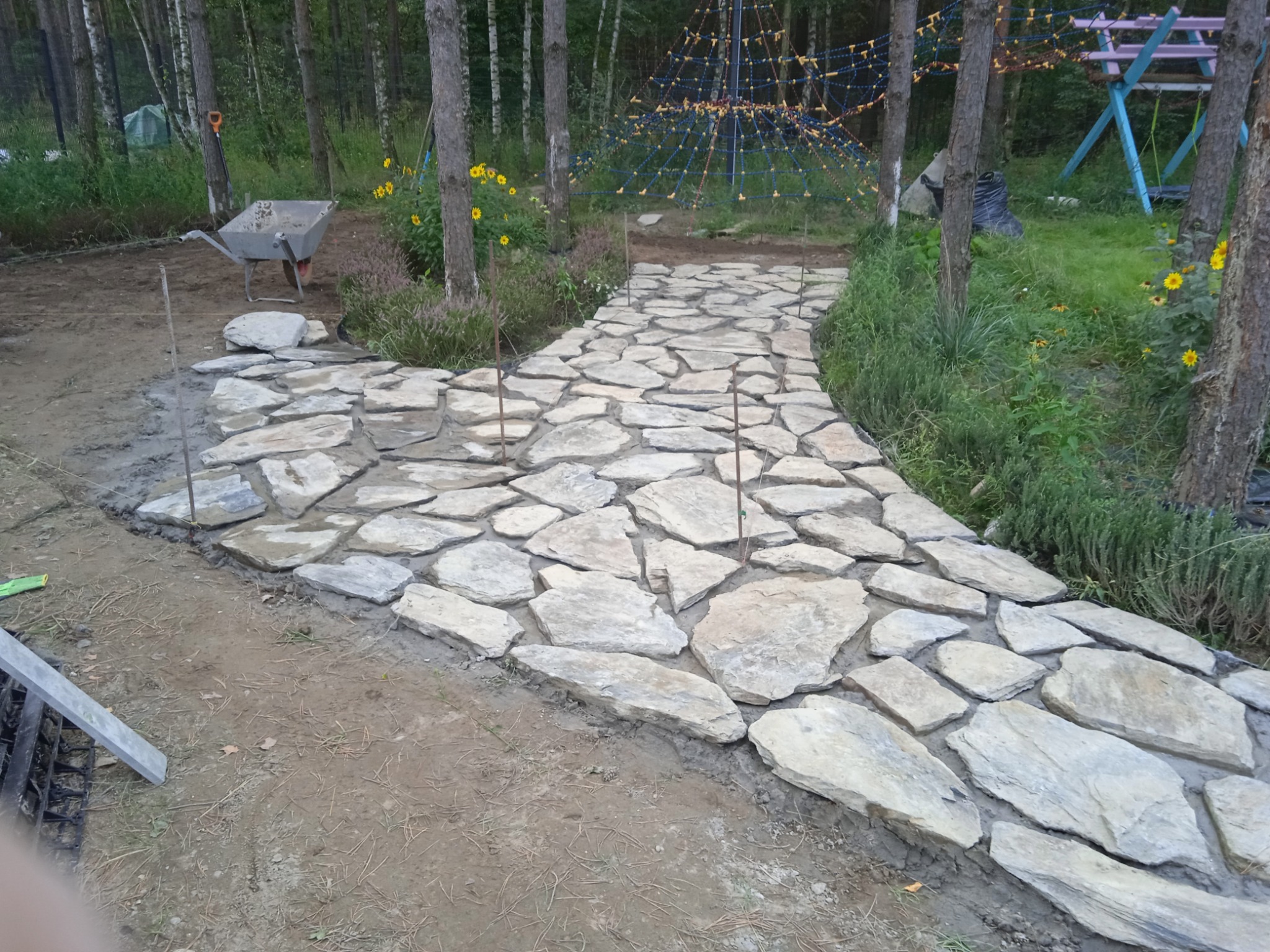 Ścieżka z nieregularnych płyt kamiennych w ogrodzie, widoczna taczka i elementy placu zabaw w tle, etap prac wykończeniowych.