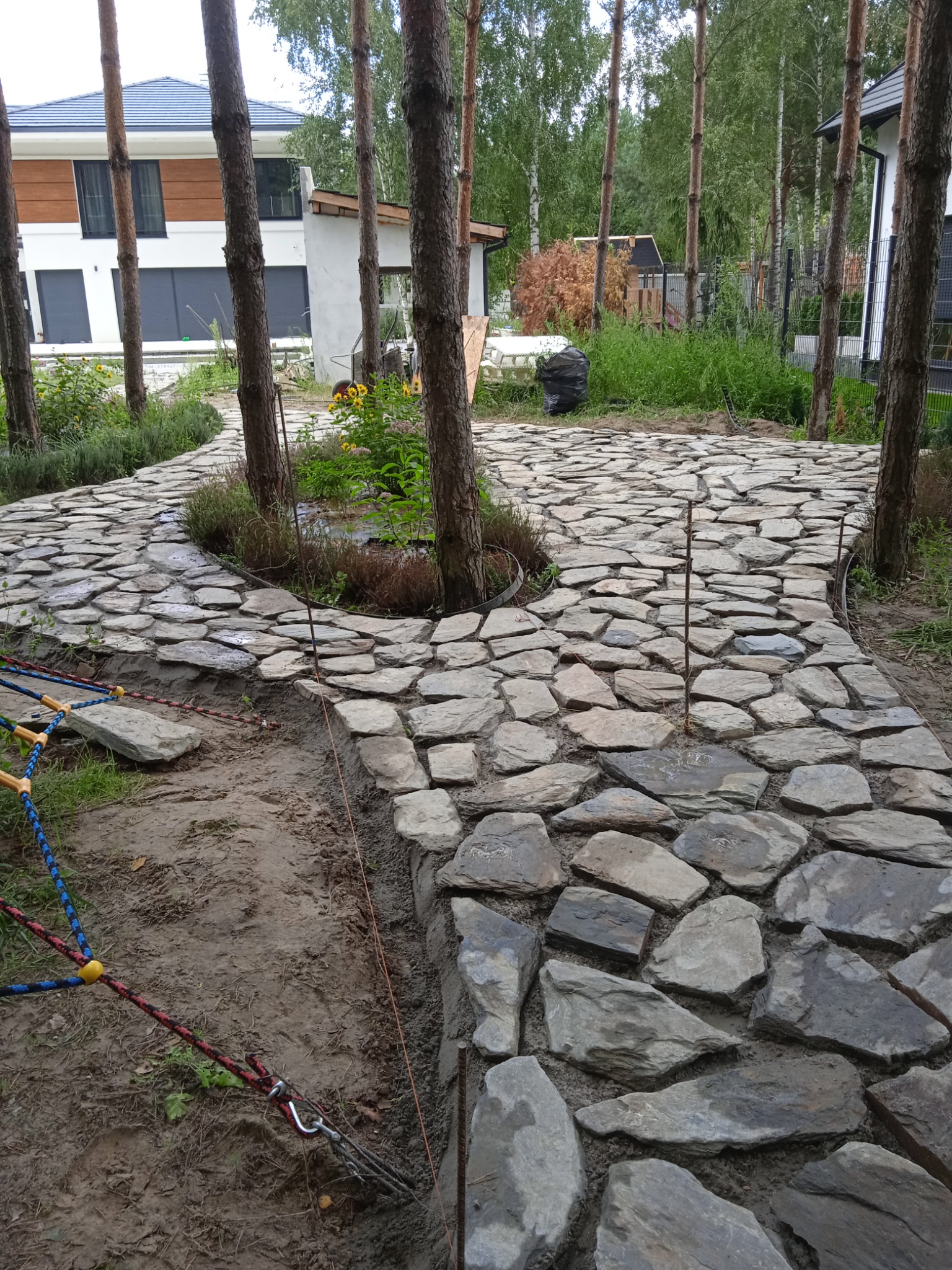 Ścieżka z nieregularnych kamieni prowadząca przez ogród z drzewami, widoczne elementy mocujące i teren w trakcie prac.