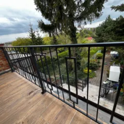 Czarna, metalowa balustrada balkonowa z geometrycznym wzorem na tle zielonego ogrodu i ceglanej ściany budynku.