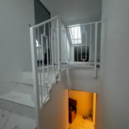 Białe, nowoczesne schody z marmurowymi stopniami i geometryczną, białą barierką, widok z góry na dół, w tle pomieszczenie z oknem dachowym.