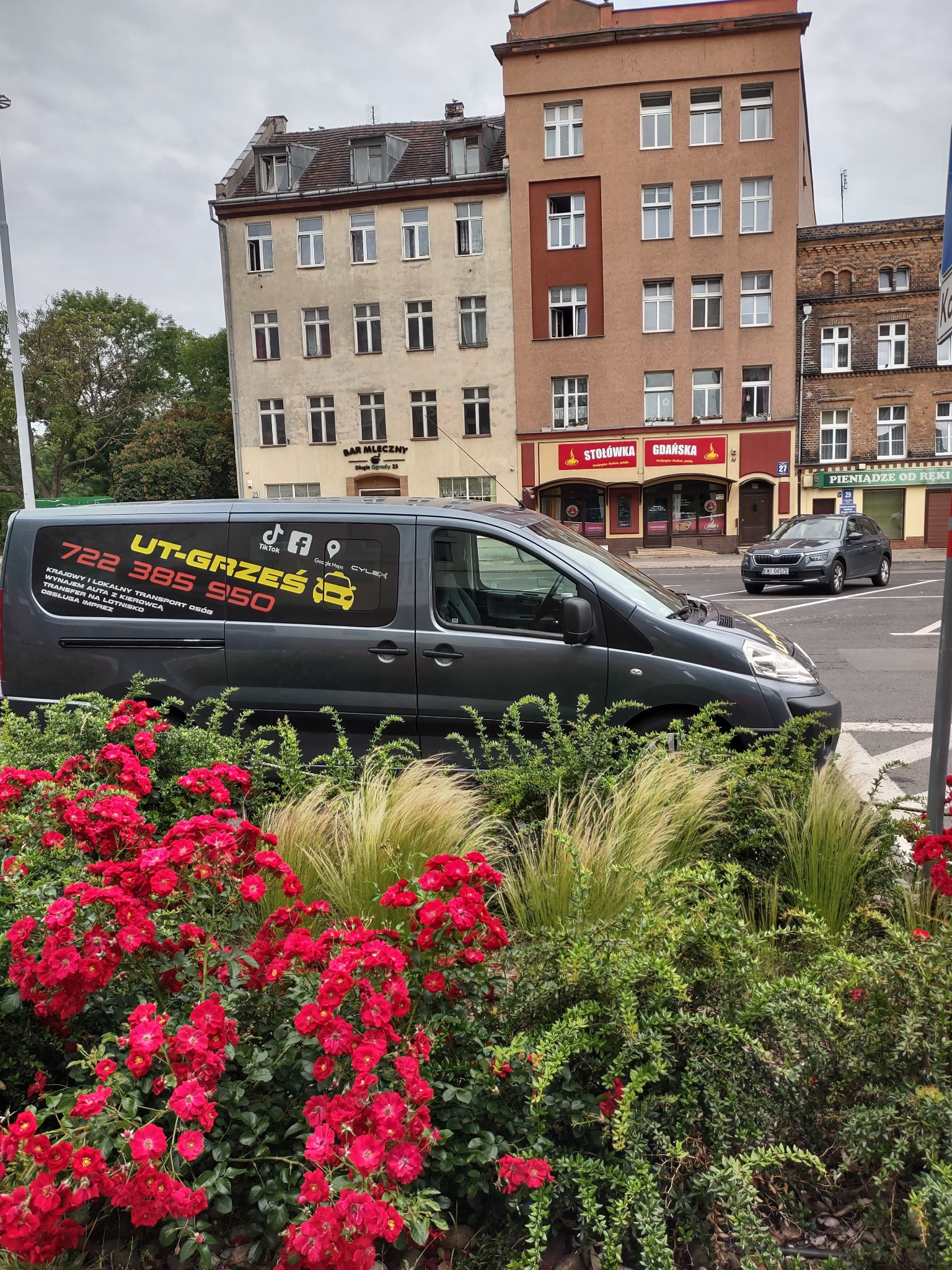 Szary van z reklamą firmy transportowej 'UT-Grześ' na tle miejskiej zabudowy i rabaty z czerwonymi różami.