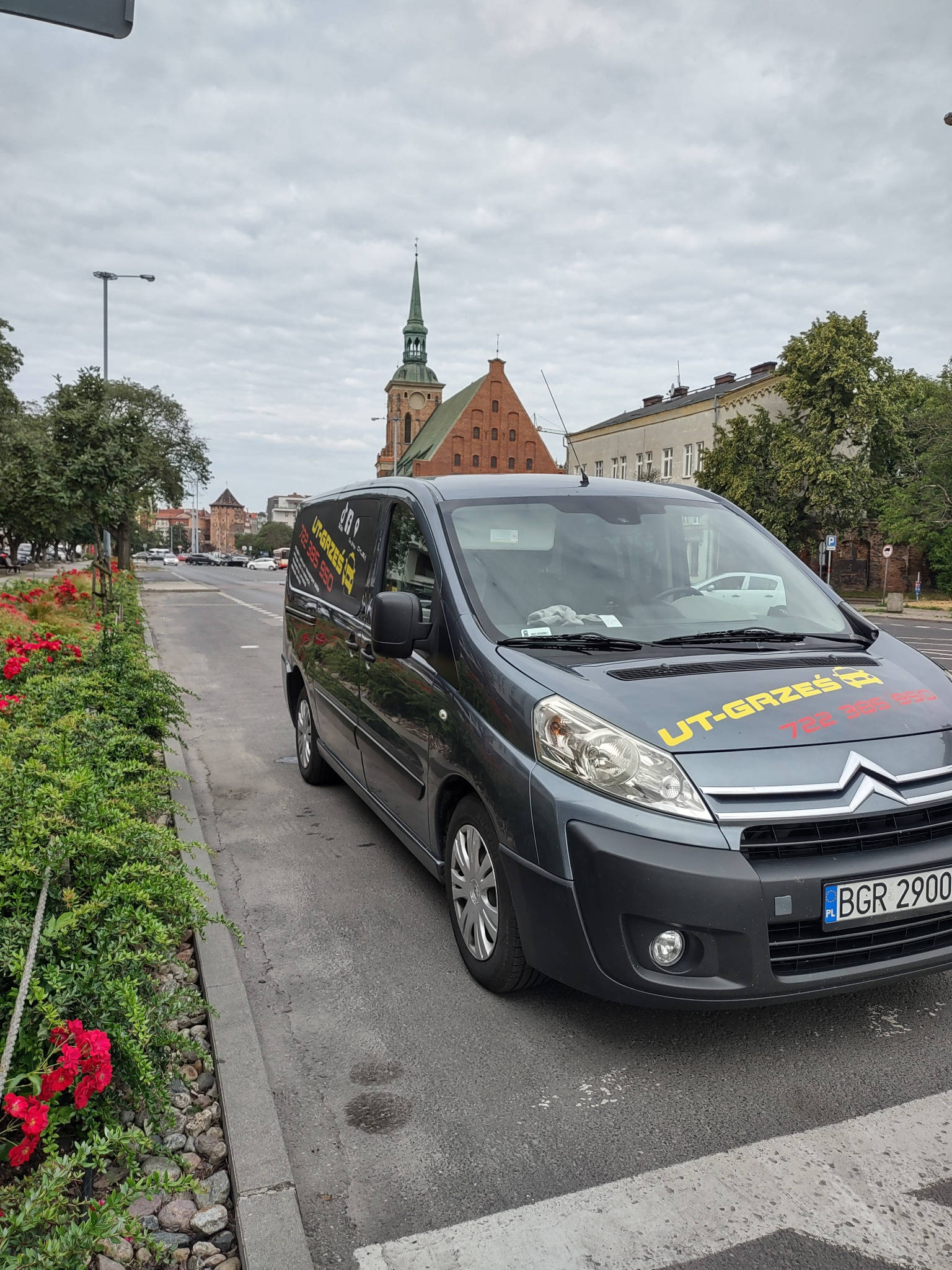 Szary samochód dostawczy firmy z żółto-czerwonym logo 'UT-GRZEŚ' zaparkowany na ulicy, z widocznym kościołem w tle i rabatą czerwonych kwiatów po lewej stronie.