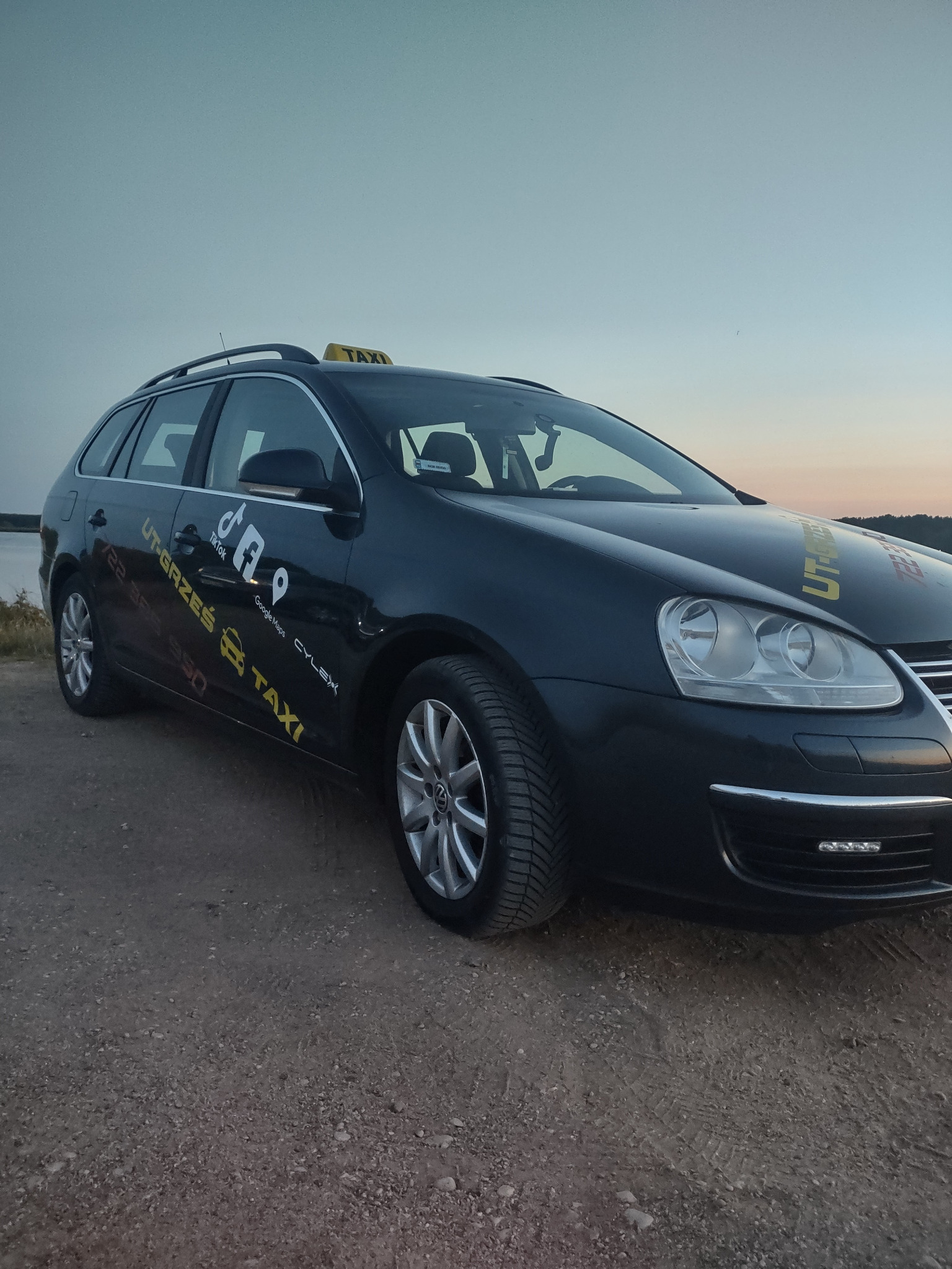 Czarny samochód kombi taxi z żółtym napisem TAXI na dachu i naklejkami z logo firmy przewozowej i social mediów na boku, zaparkowany na szutrowej drodze o zmierzchu.