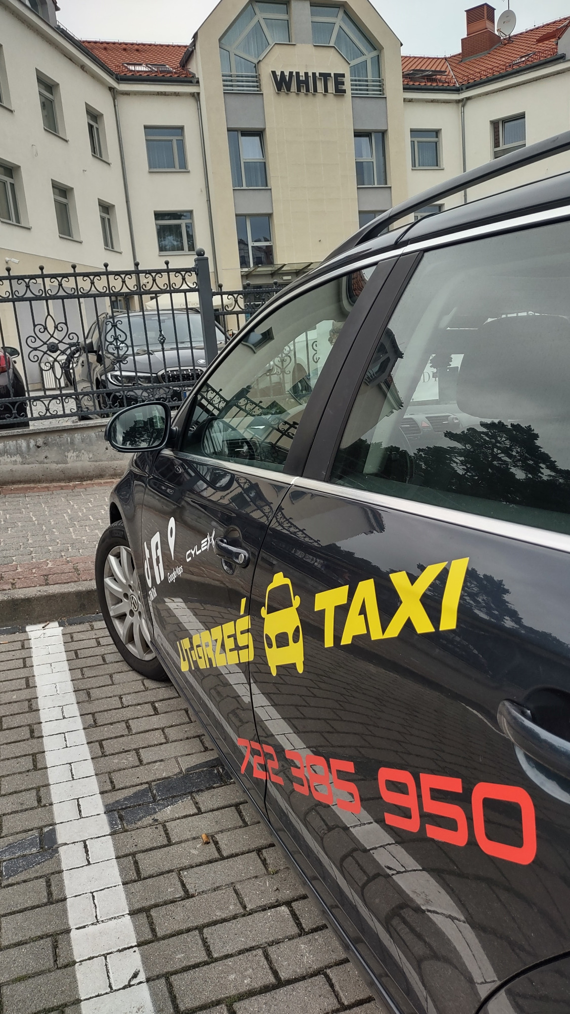 Czarny samochód osobowy z żółtym napisem 'TAXI' i ikoną taksówki na tle budynku 'WHITE', zaparkowany na parkingu z kostki brukowej.