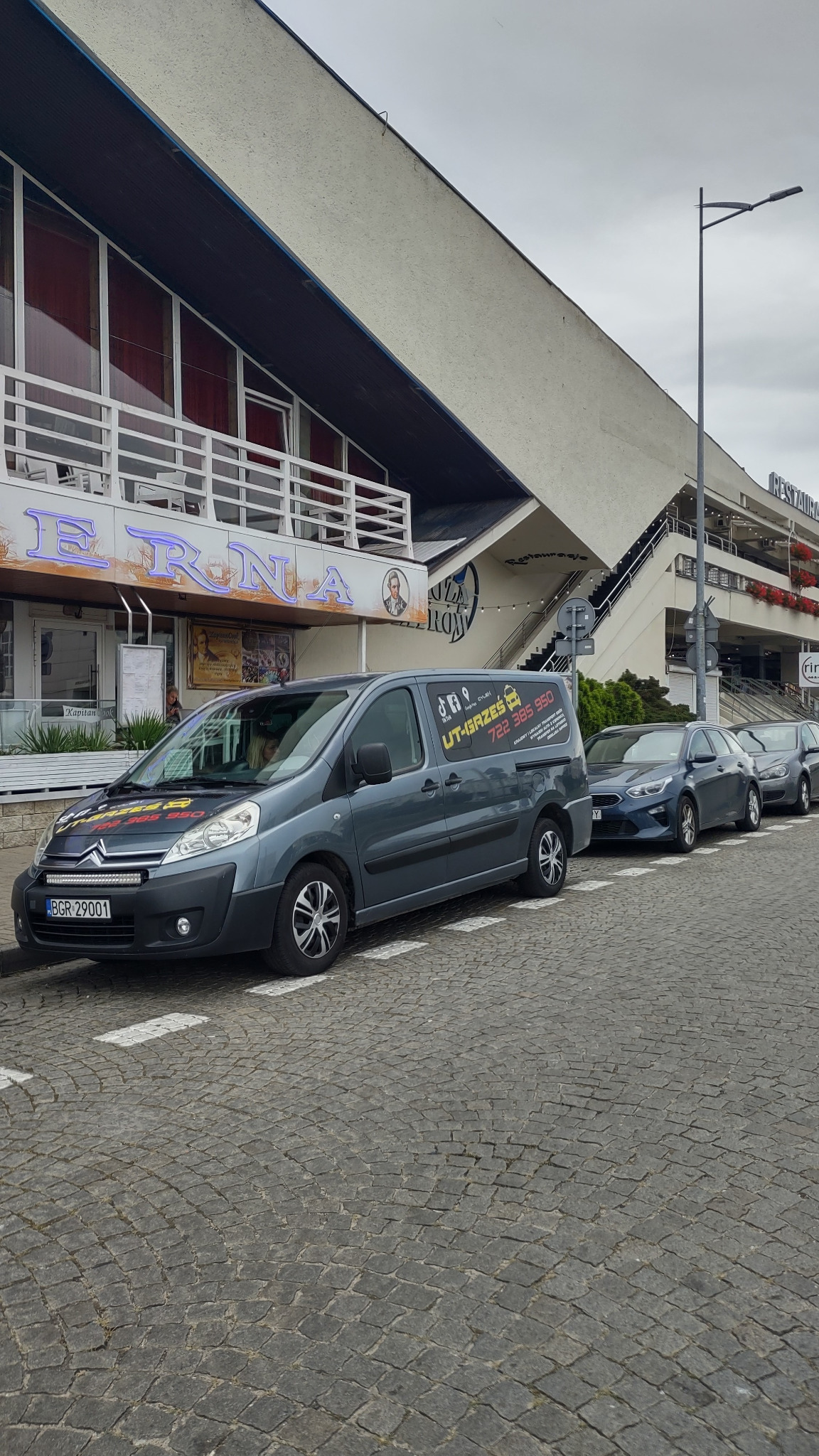 Szary samochód dostawczy z naklejkami firmy transportowej 'UT-GRZEŚ' zaparkowany na brukowanej ulicy przed restauracją 'ERNA', w tle budynek z schodami.