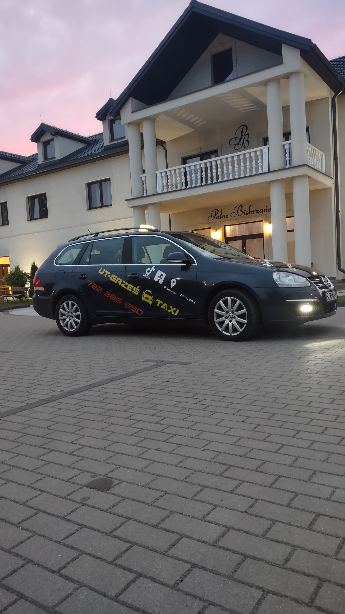 Ciemny samochód typu kombi z oznaczeniami Taxi, zaparkowany przed budynkiem z kolumnami i napisem 'Pałac Biebrzański' o zmierzchu.