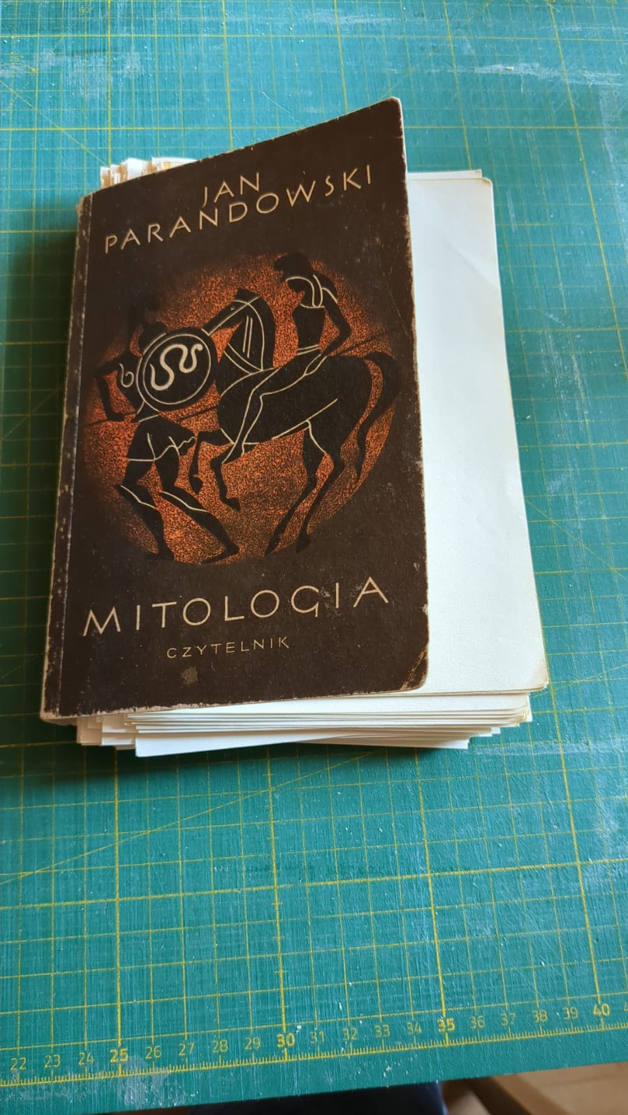 Stara książka 'Mitologia' Jan Parandowski, z widocznymi śladami użytkowania, leży na stosie białych kartek na zielonej macie introligatorskiej.