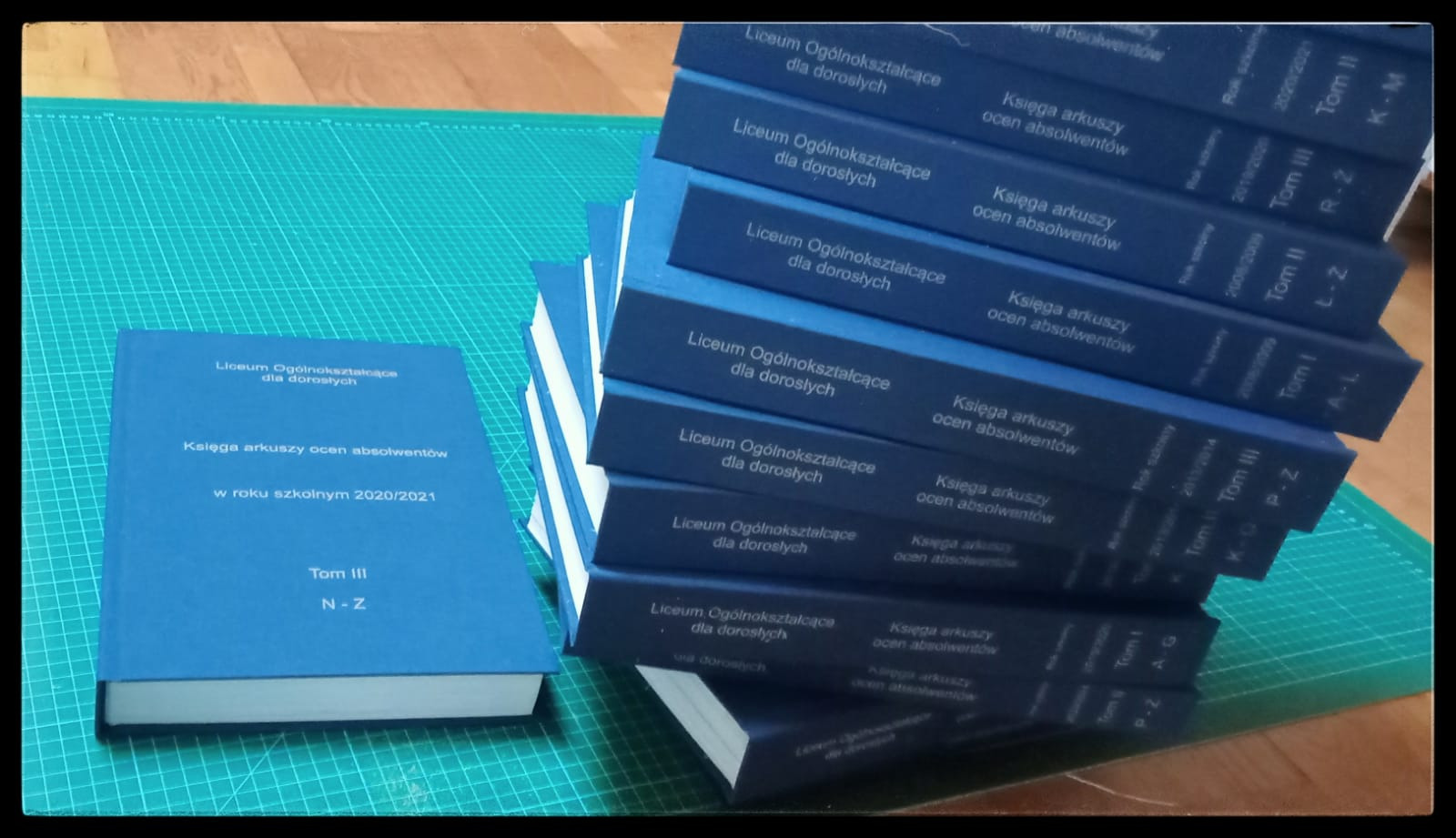 Stos niebieskich, oprawionych ksiąg 'Księga arkuszy ocen absolwentów Liceum Ogólnokształcącego dla dorosłych', jedna leży obok. Widok z góry, na zielonej macie.