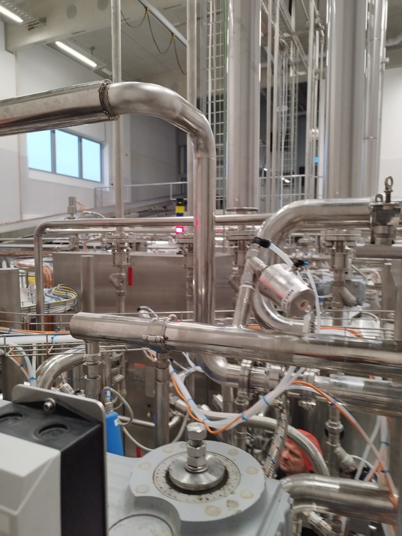 Skonstruowany system rur ze stali nierdzewnej w hali produkcyjnej, widoczny pracownik w czerwonym kasku w tle.