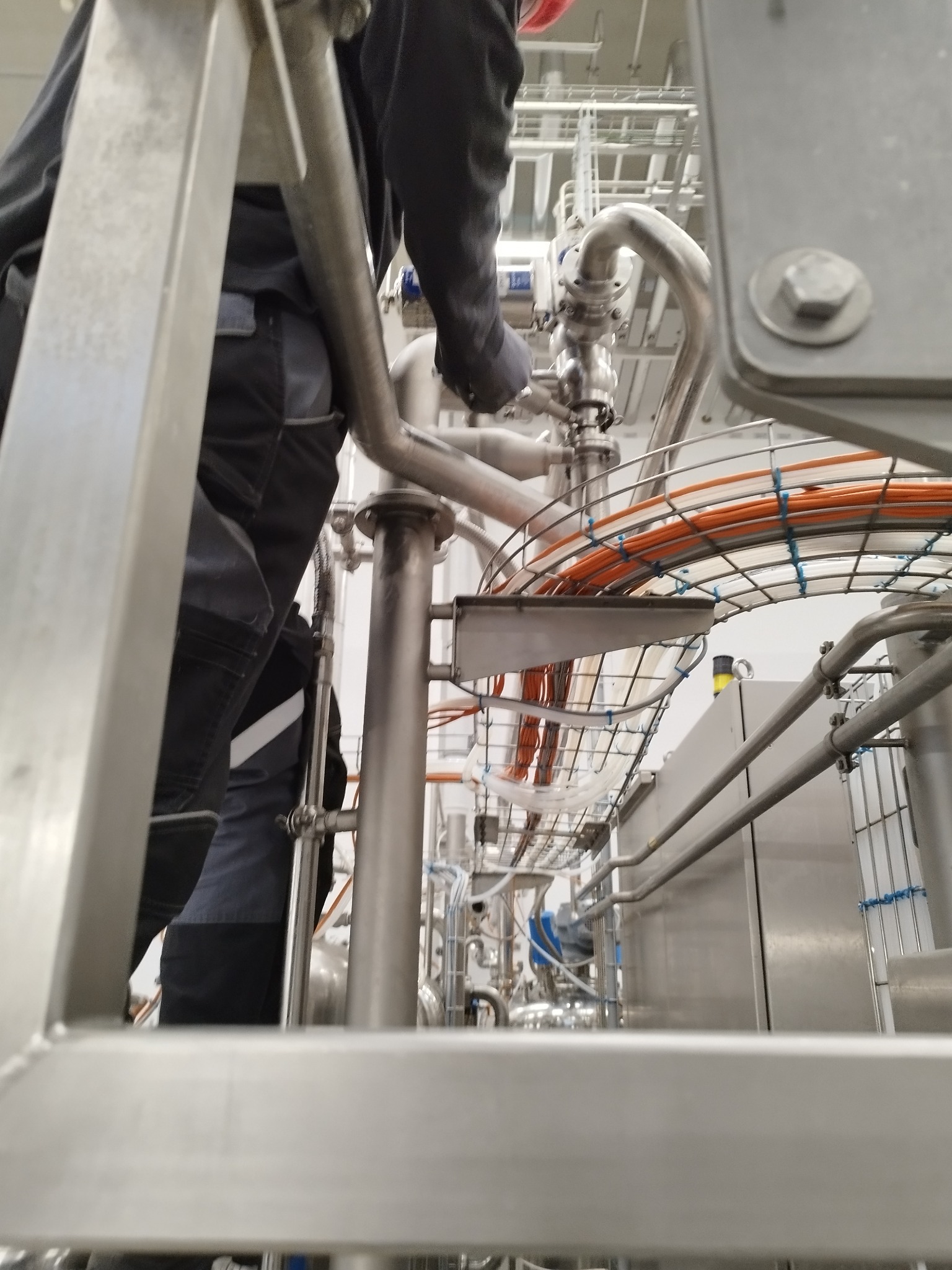 Pracownik w kombinezonie roboczym łączy elementy instalacji przemysłowej, widoczne rury ze stali nierdzewnej i okablowanie w metalowej klatce.