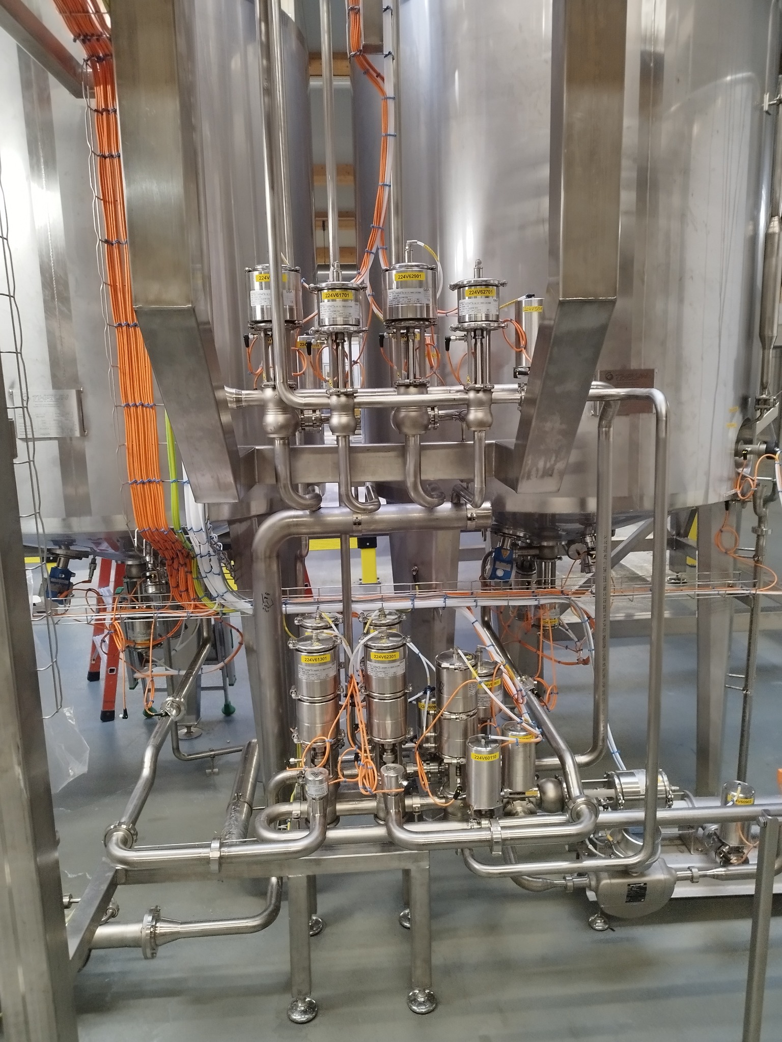 Fragment instalacji przemysłowej ze stali nierdzewnej z widocznymi zbiornikami, rurami, zaworami i okablowaniem w kolorze pomarańczowym, umieszczonej na szarej posadzce.