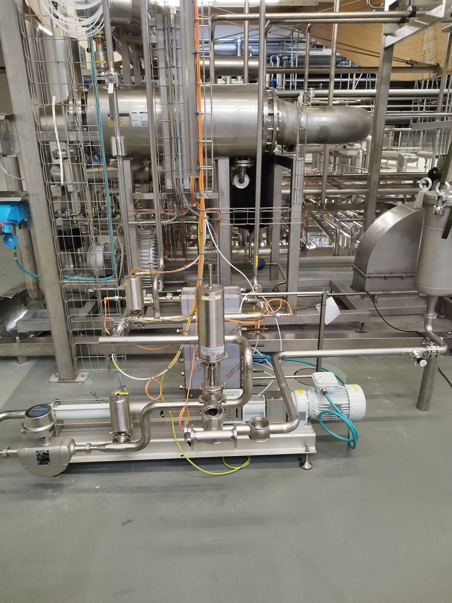 Kompleksowa instalacja przemysłowa ze stali nierdzewnej z licznymi rurami, zbiornikami i silnikami, widoczne okablowanie i elementy sterujące, na gładkiej, szarej posadzce.