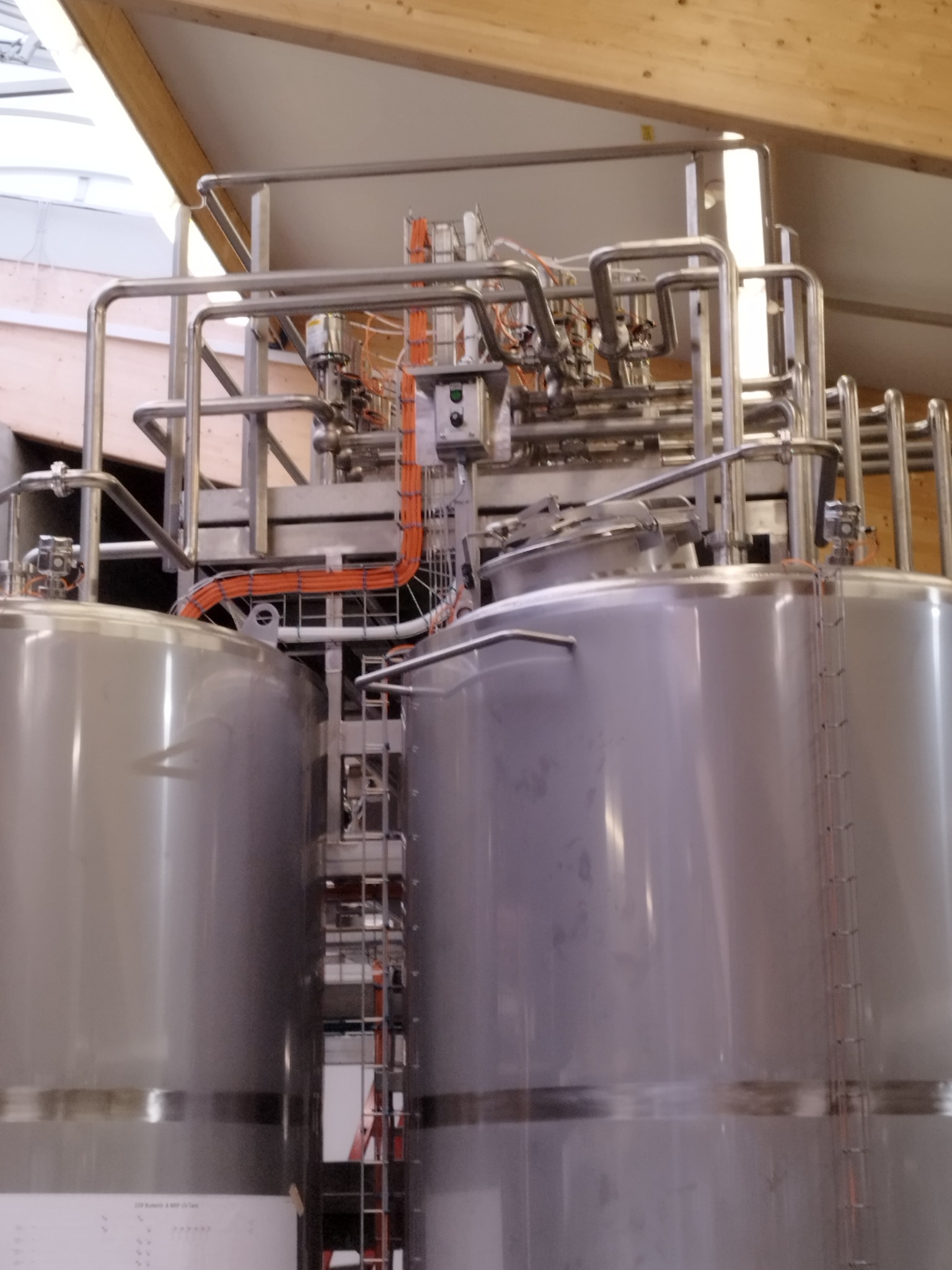 Dwa duże, srebrne zbiorniki przemysłowe z rurami i przewodami, umieszczone w hali z drewnianą konstrukcją stropu.