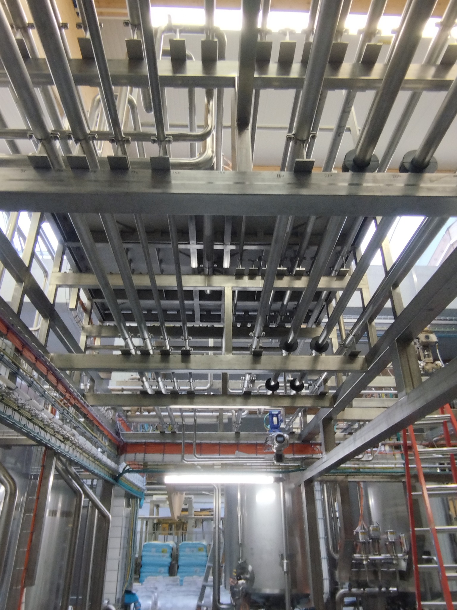 Skomplikowana instalacja rur i konstrukcji stalowych w hali produkcyjnej, widok od dołu, z widocznymi zbiornikami i elementami wyposażenia.