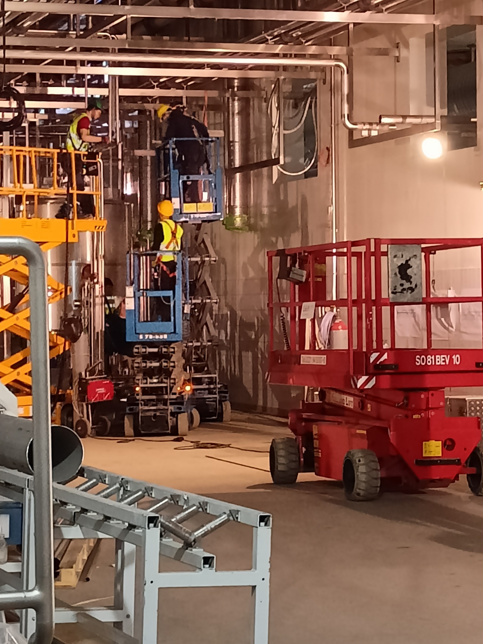 Montaż instalacji przemysłowej: spawacze na podnośnikach koszowych pracują przy rurociągach w hali, widoczny fragment konstrukcji stalowej i podajnik rur.