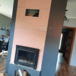 Obudowa kominka w trakcie montażu z płyt kartonowo-gipsowych, widoczne otwory montażowe i rama wkładu kominkowego. W tle fragment jadalni i drzwi.