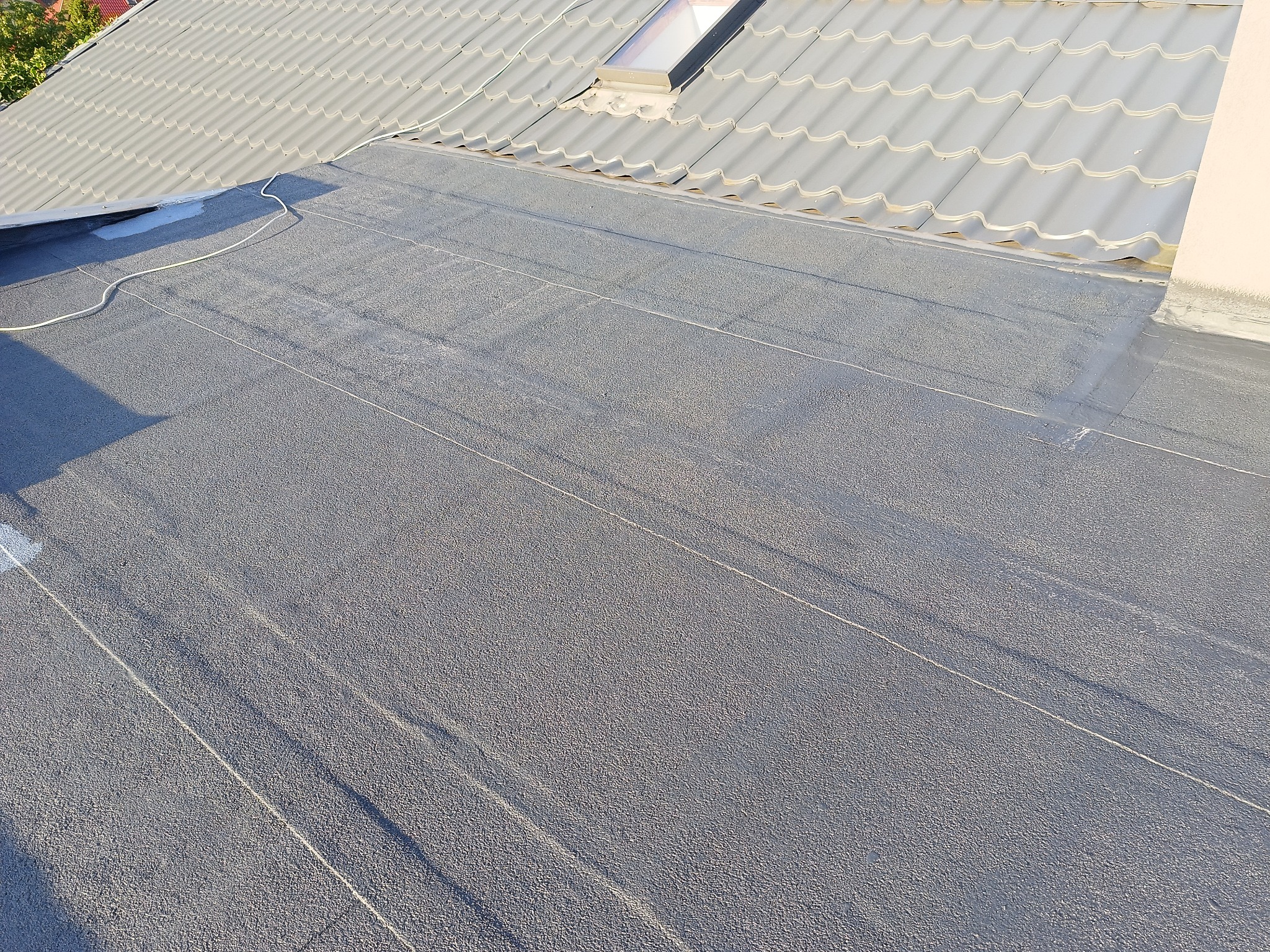 Szary, płaski dach pokryty papą termozgrzewalną, widoczne łączenia, fragment dachu z dachówki ceramicznej w tle, kabel biegnący po powierzchni dachu, element okna dachowego.