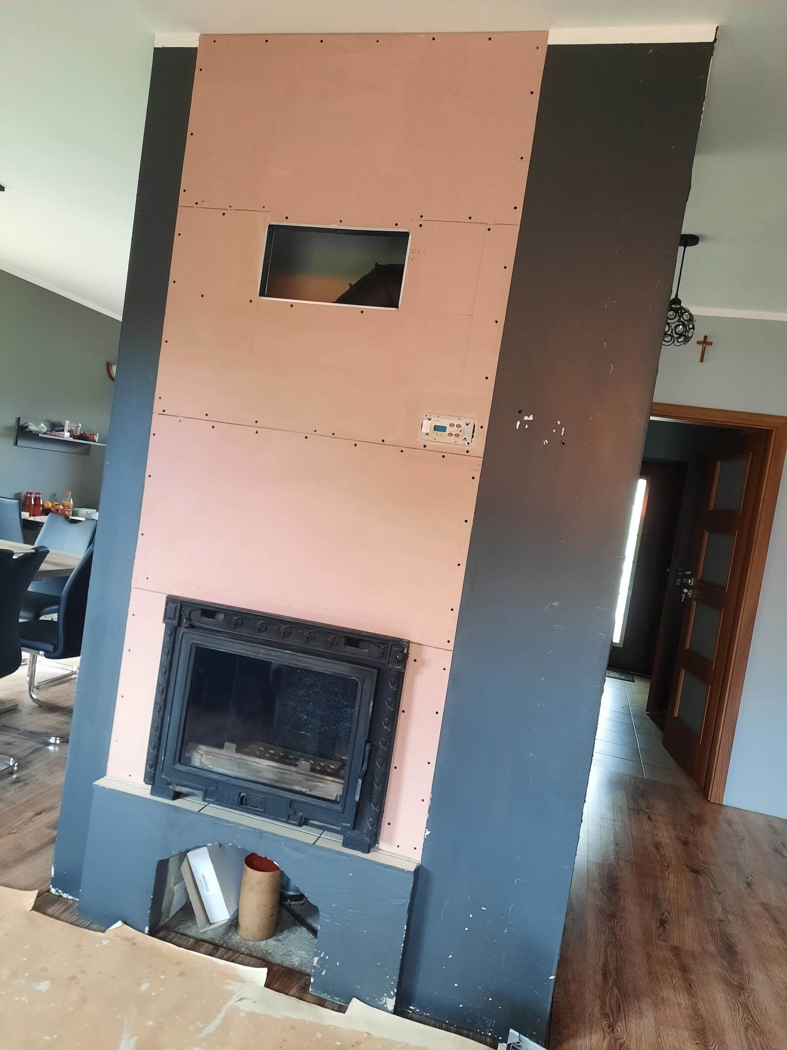 Obudowa kominka w trakcie montażu z płyt kartonowo-gipsowych, widoczne otwory montażowe i rama wkładu kominkowego. W tle fragment jadalni i drzwi.