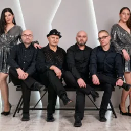 Zespół muzyczny: dwóch wokalistów w błyszczących sukienkach i trzech muzyków w czarnych strojach pozuje na tle białej ściany z geometrycznymi wzorami.