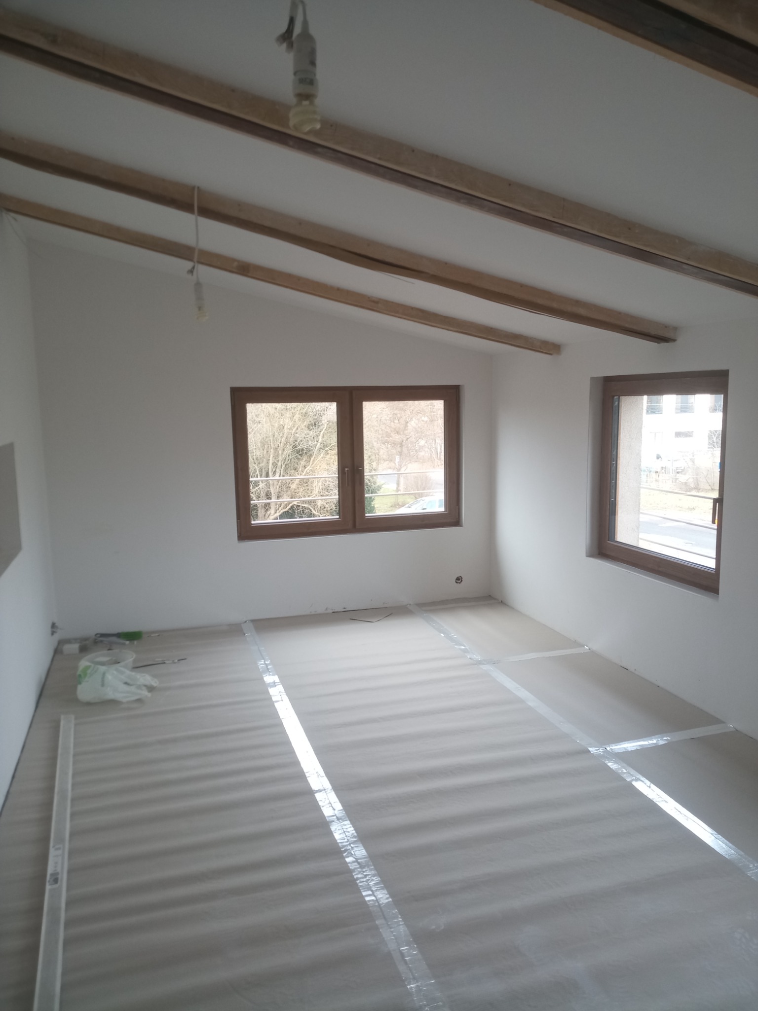 Pokój w trakcie przygotowania do prac wykończeniowych, podłoga zabezpieczona folią i taśmą, widoczne drewniane belki pod sufitem, dwa okna z widokiem na zewnątrz.