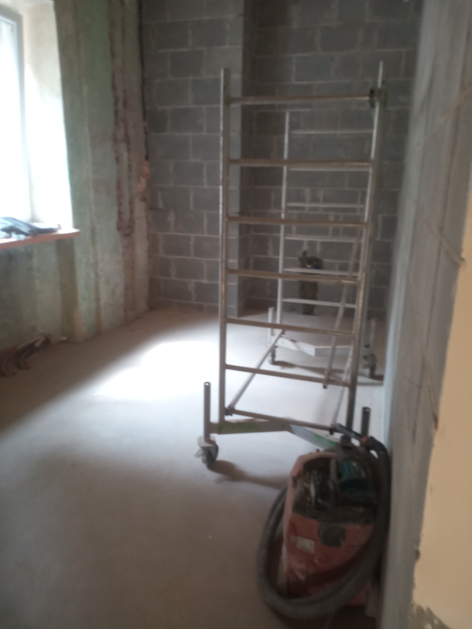 Wnętrze w trakcie remontu: surowe ściany z betonu komórkowego, metalowe rusztowanie na kółkach i przemysłowy odkurzacz na podłodze.