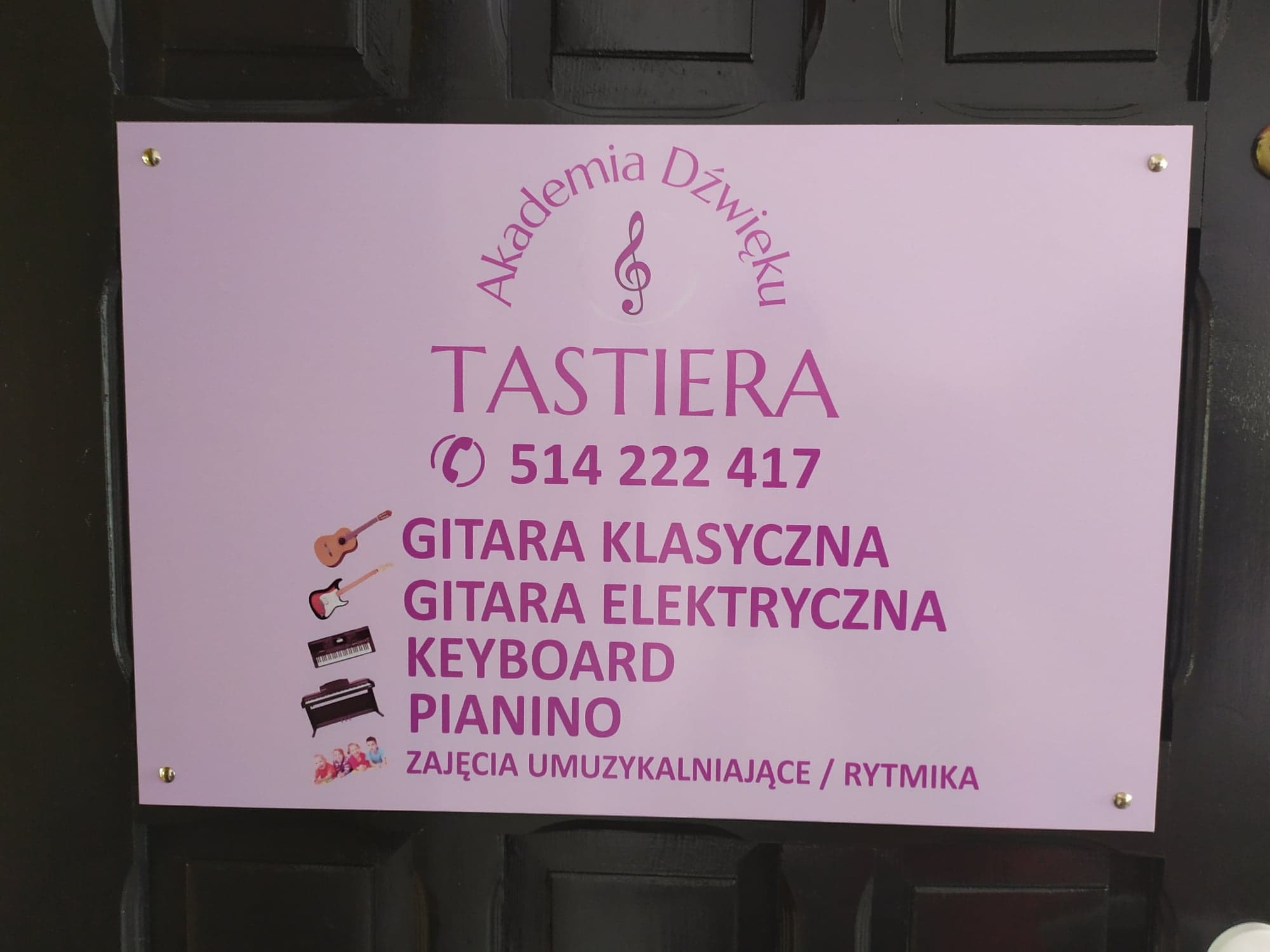 Fioletowa tabliczka informacyjna przytwierdzona do ciemnych drzwi, z logo Akademii Dźwięku TASTIERA, numerem telefonu i listą oferowanych zajęć: gitara klasyczna, gitara elektryczna, keyboard...