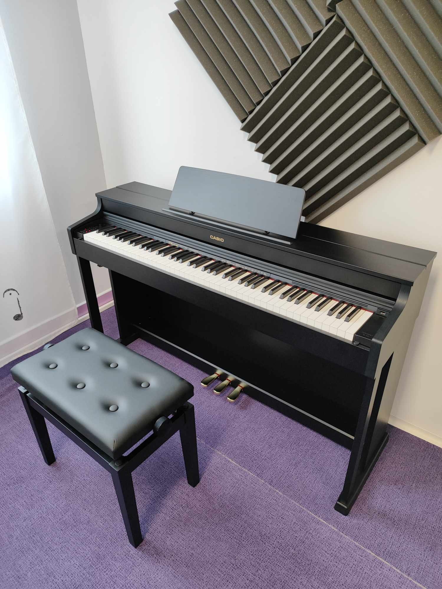 Czarne pianino cyfrowe Casio z ławką na fioletowym dywanie, na ścianie panele akustyczne.