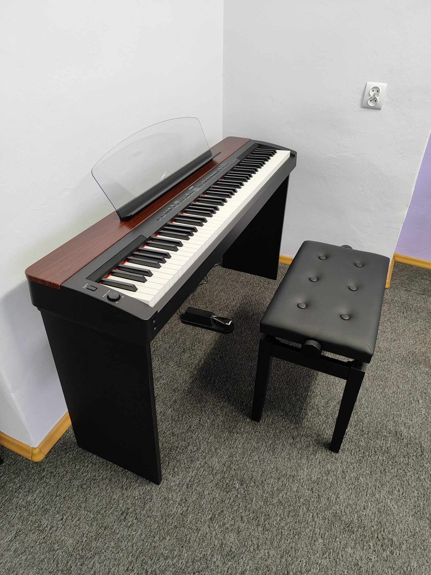 Cyfrowe pianino z mahoniową obudową i czarną podstawą, ustawione obok czarnego, tapicerowanego stołka na szarym dywanie.