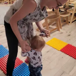 Kobieta w fartuchu z motywem jeleni trzyma za ręce małe dziecko chodzące po kolorowych, sensorycznych matach podłogowych w pomieszczeniu z drewnianymi meblami.
