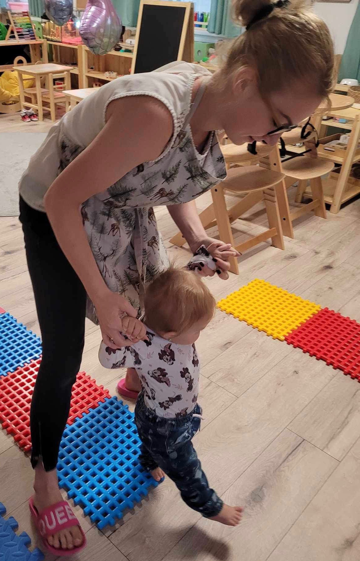 Kobieta w fartuchu z motywem jeleni trzyma za ręce małe dziecko chodzące po kolorowych, sensorycznych matach podłogowych w pomieszczeniu z drewnianymi meblami.
