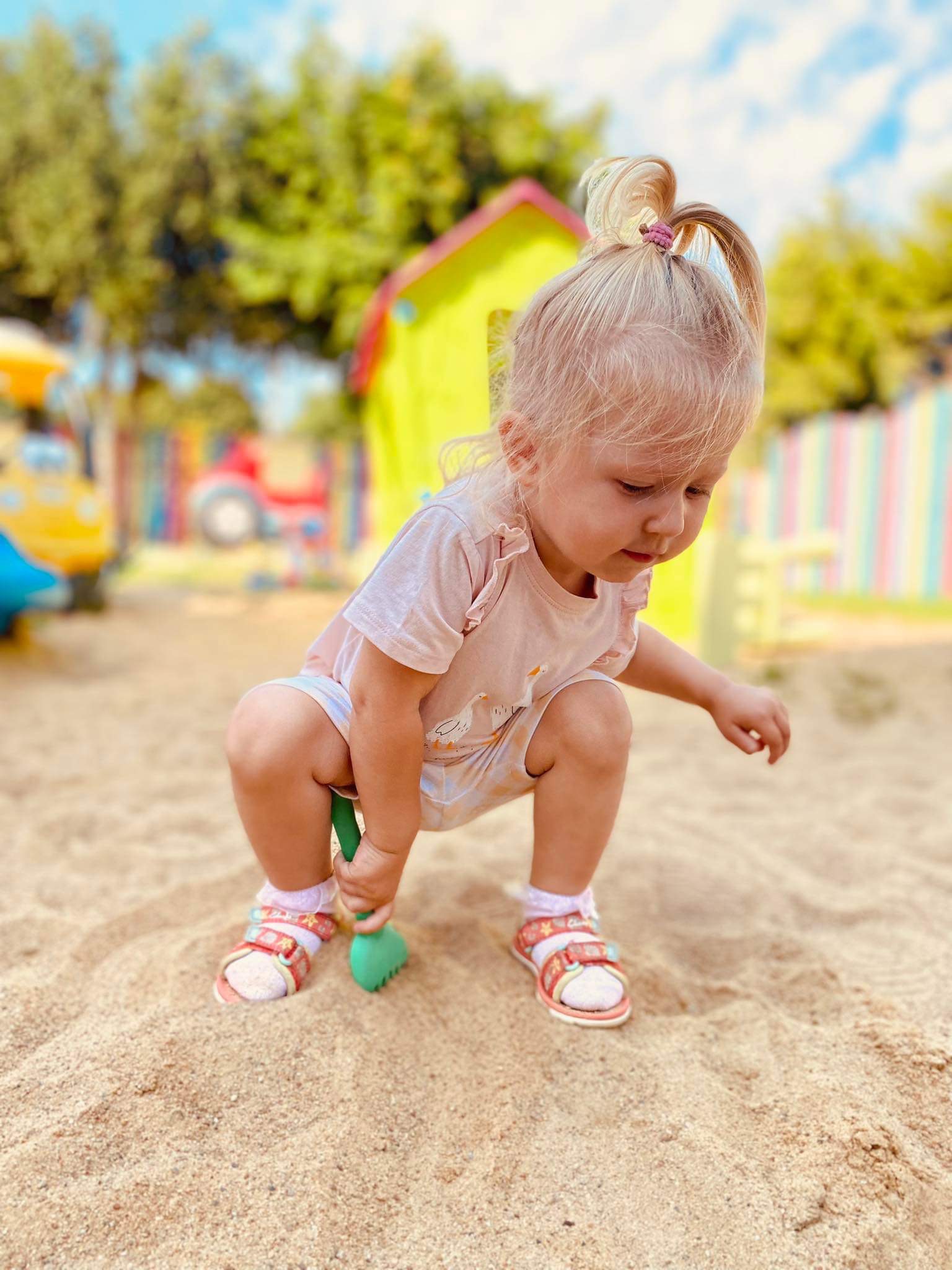 Dziewczynka w różowej koszulce i sandałach bawi się zieloną łopatką w piasku na placu zabaw z kolorowym domkiem w tle.
