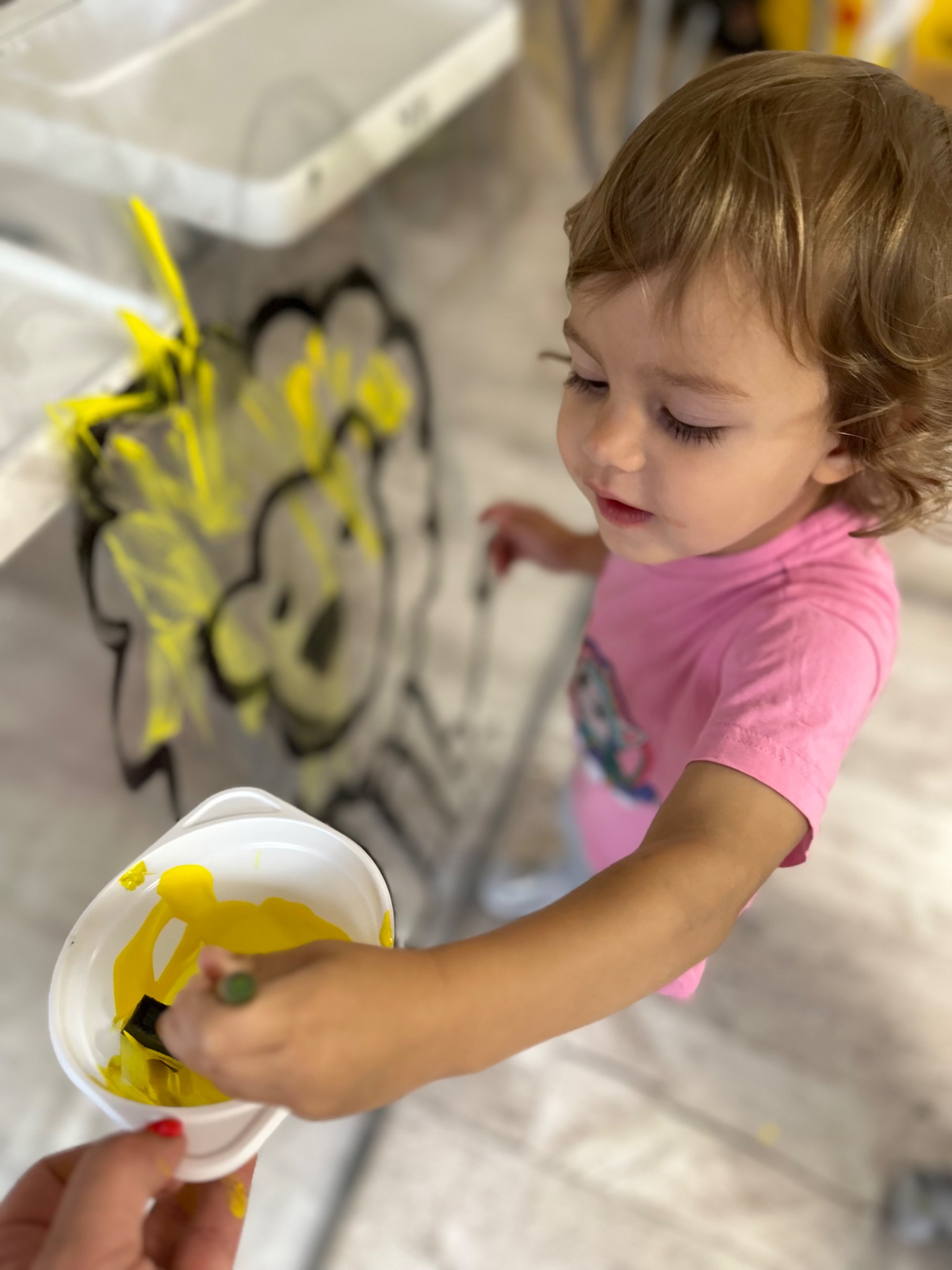 Dziewczynka w różowej koszulce maluje żółtą farbą po pionowej powierzchni, obok dłoń dorosłego trzymająca pojemnik z farbą i pędzlem.