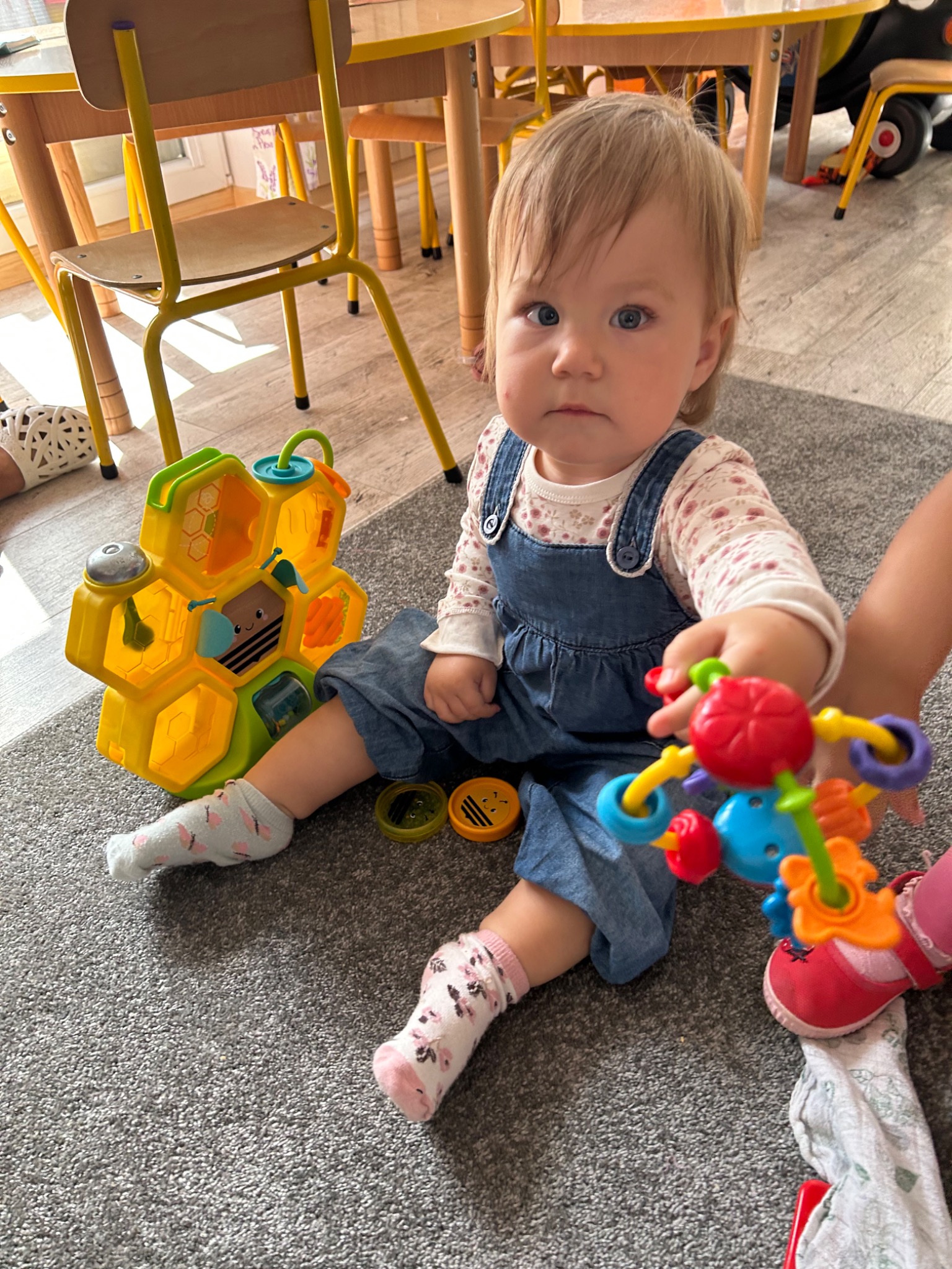 Dziewczynka w dżinsowym ogrodniczce siedzi na szarym dywanie i bawi się kolorowymi zabawkami, w tle widoczne żółte krzesełka i stoliki dziecięce.