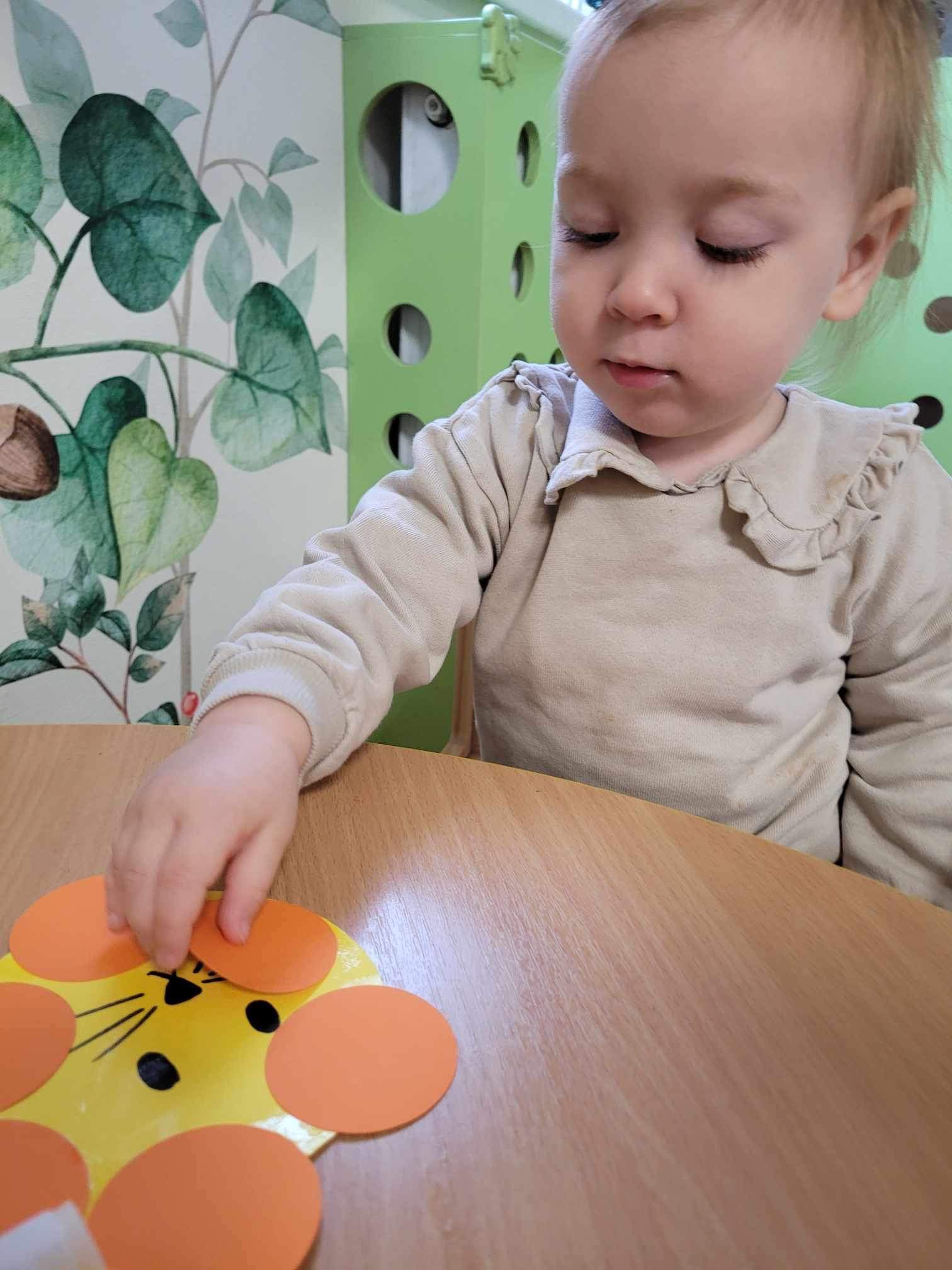 Mała dziewczynka w beżowej bluzce siedzi przy drewnianym stole i układa pomarańczowe kółka na żółtej kartce z narysowanym lwem.