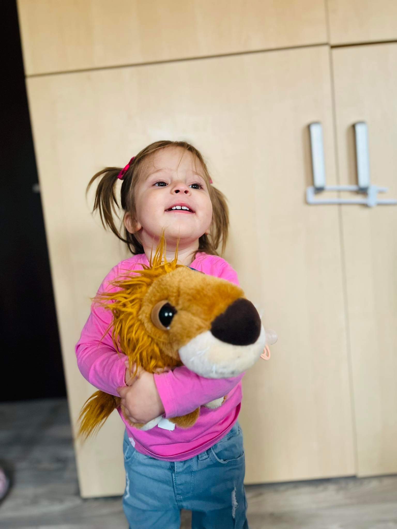 Uśmiechnięta dziewczynka w różowej bluzce i dżinsach przytula pluszowego lwa na tle jasnych szafek.