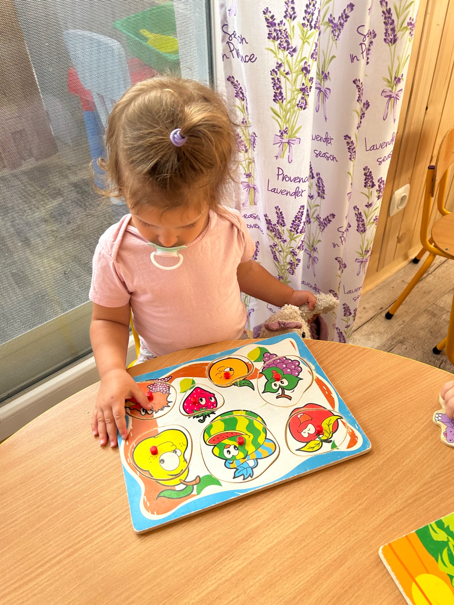 Dziewczynka z blond włosami spiętymi fioletową gumką, ubrana w różową koszulkę, siedzi przy drewnianym stole i układa drewniane puzzle z rysunkami owoców i warzyw. W tle zasłona w lawendowy wzór.