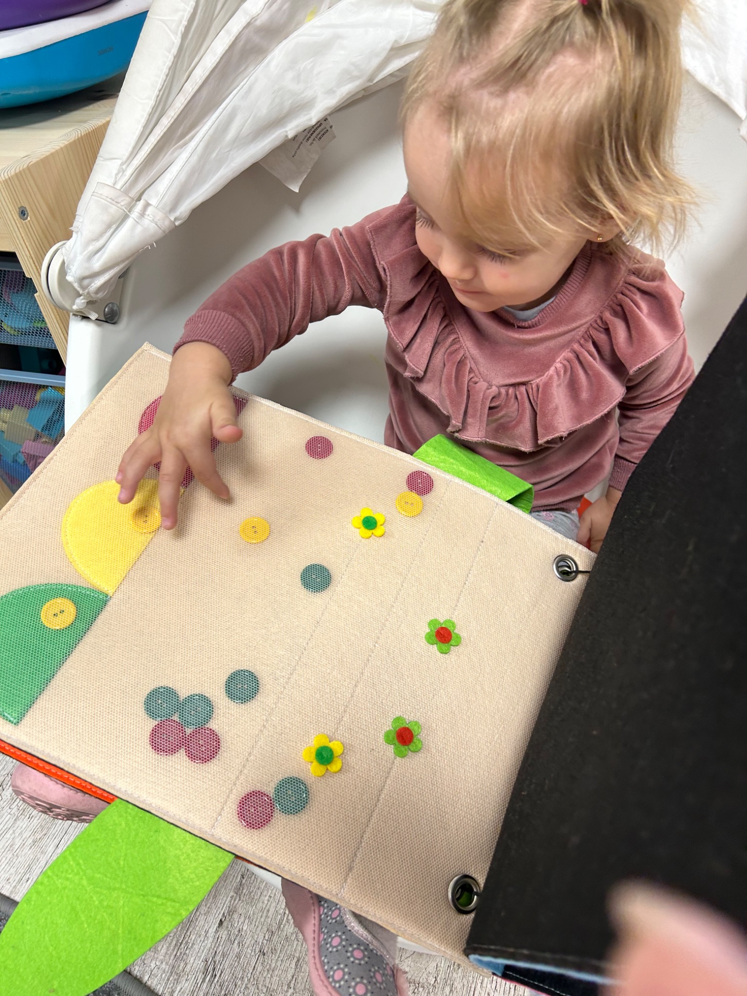 Dziewczynka w różowym welurowym ubranku bawi się na macie edukacyjnej z kolorowymi kształtami i guzikami, siedząc w białym krzesełku.