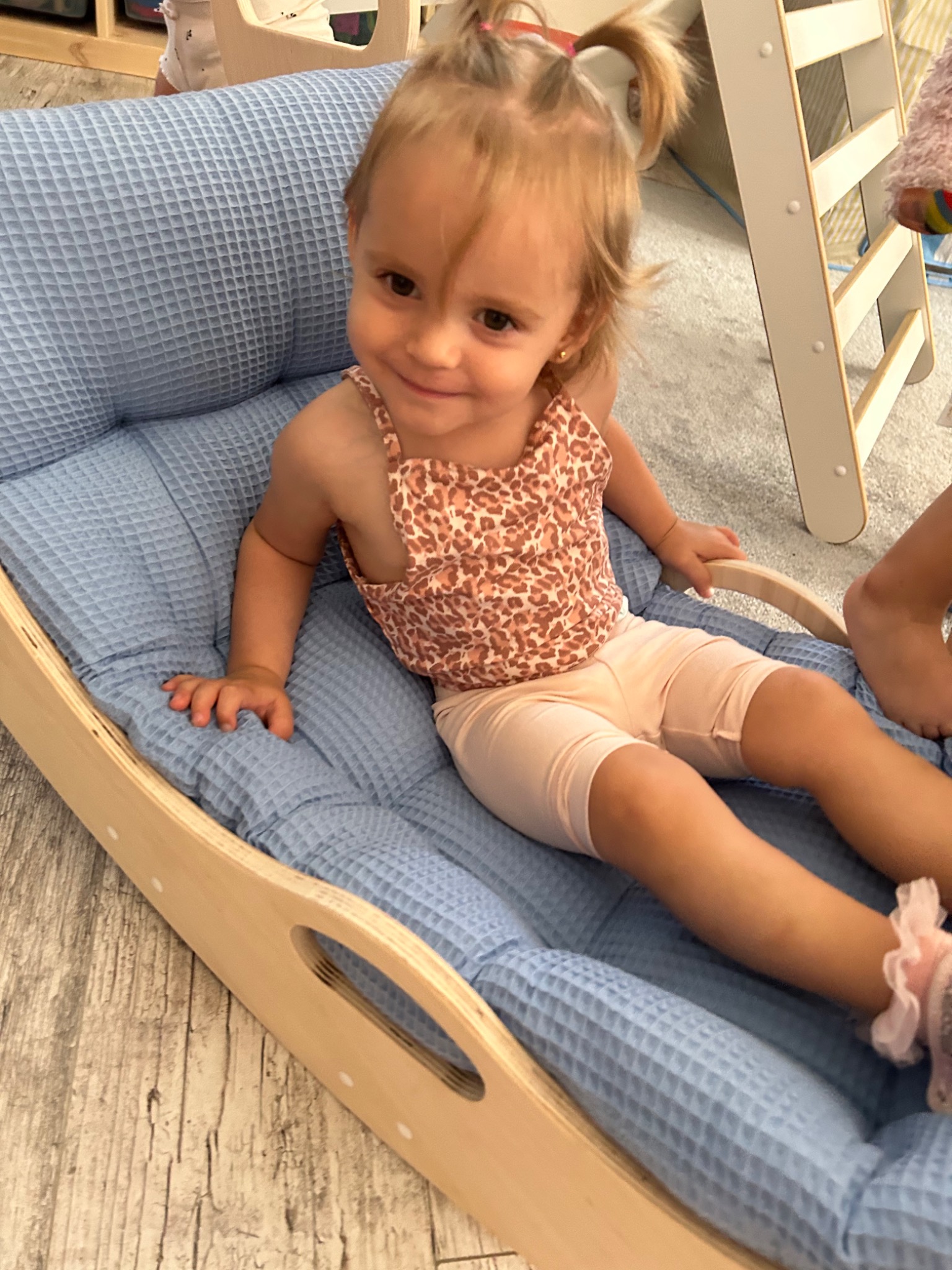 Uśmiechnięta dziewczynka siedzi w drewnianym bujaku z niebieską, pikowaną poduszką, obok drabinka wspinaczkowa.